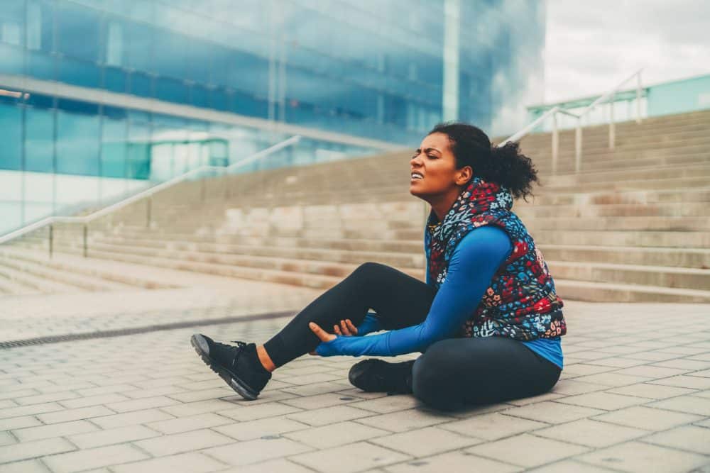 A woman sits on the ground outside of a building holding her leg in pain.