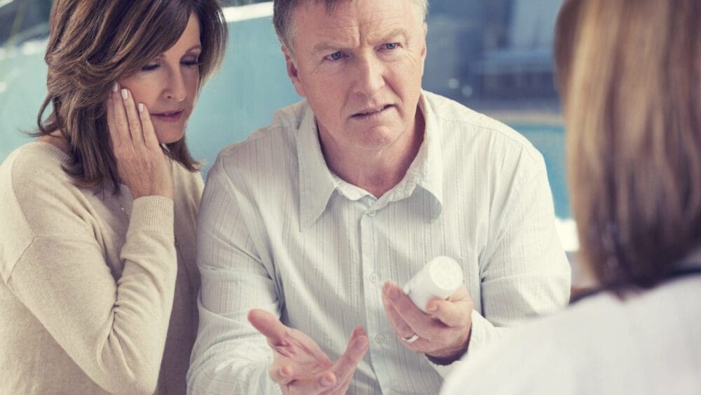 A mature couple speaks to their pharmacist about prescription drugs as the wife holds her jaw in pain.