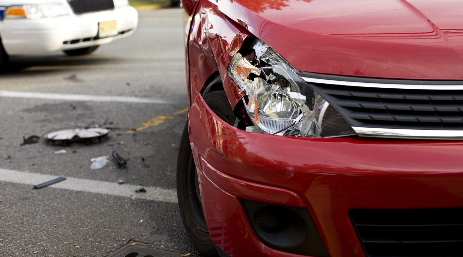 The front of a damaged red car, pieces of a car on the ground, and a white car in the background indicating a recent collision.