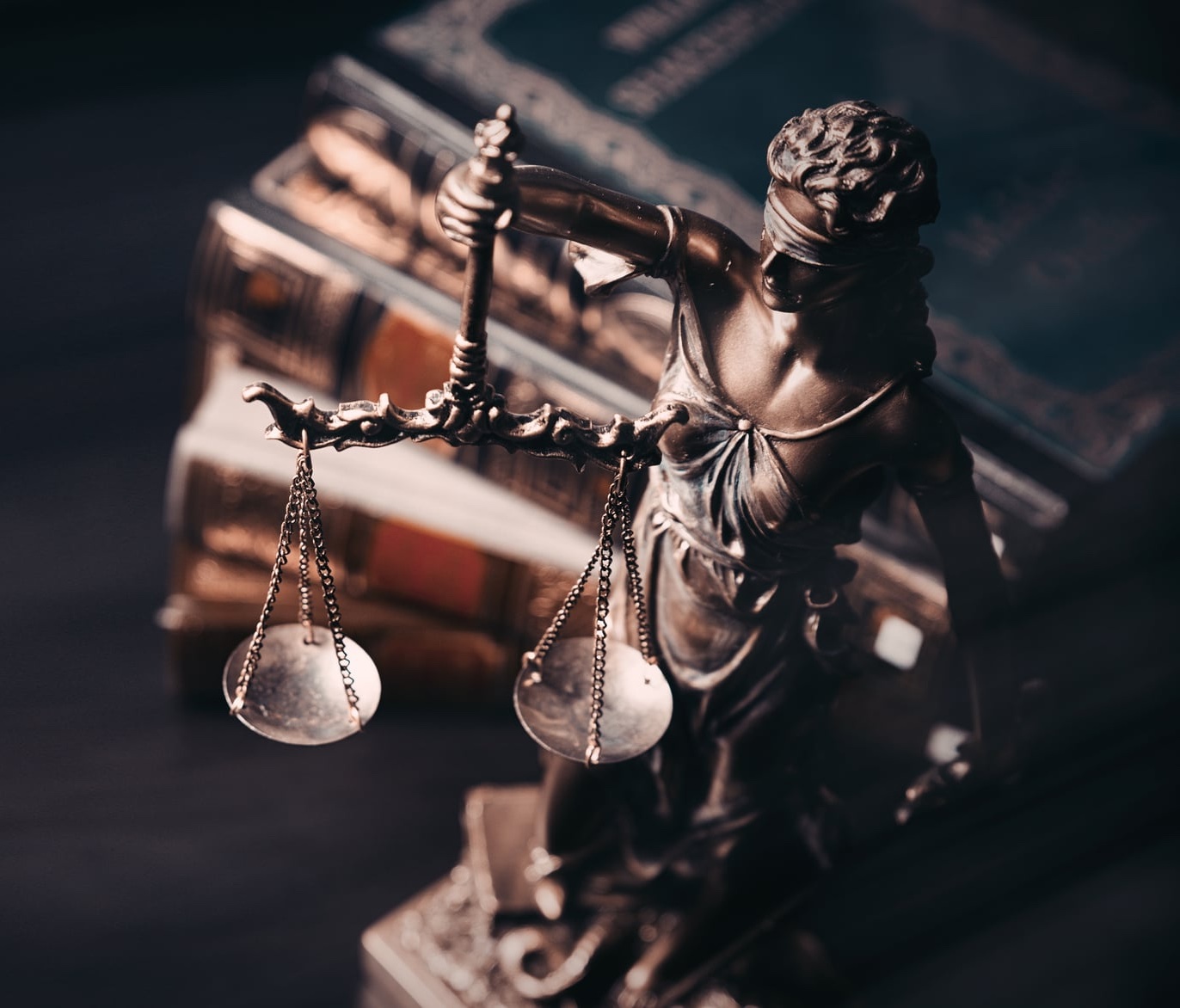 A bronze statue of Lady Justice holding scales stands in front of a stack of historic legal books.