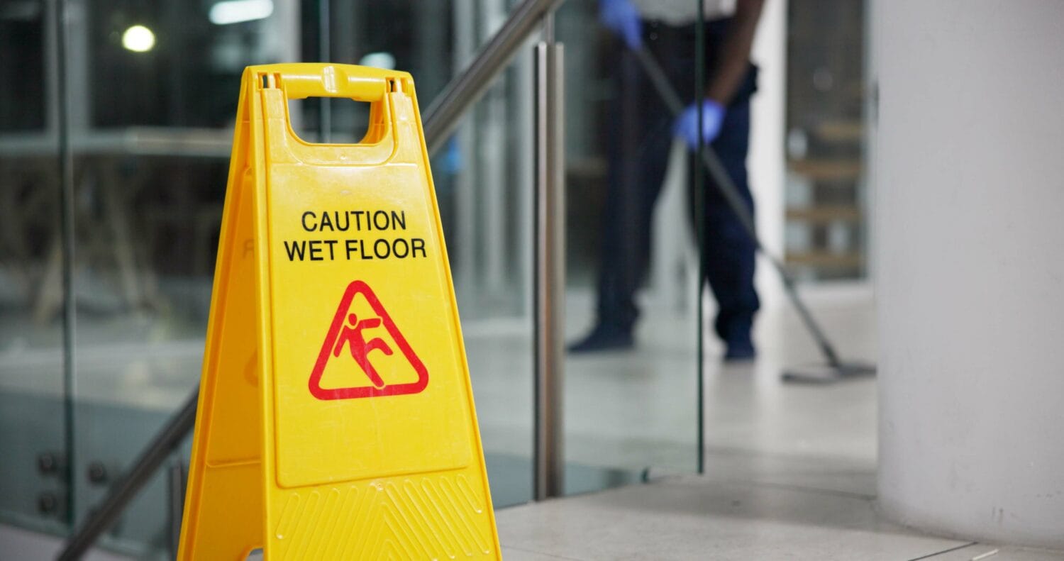 A wet floor sign is posted near a staircase in a commercial building as a janitor mops the floor in the background