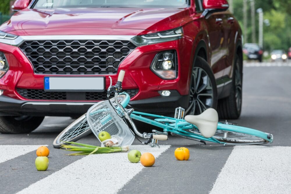 A red SUV is stopped at a crosswalk where a blue bicycle lies on the ground with produce spilling out of its basket.