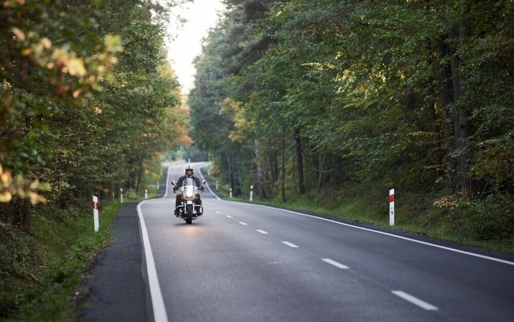 A motorcyclist in black leather riding gear rides down a rural road surrounded by trees.