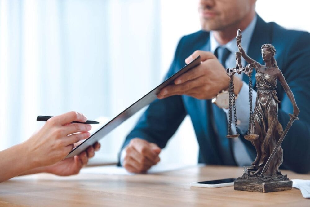 An attorney holds a clipboard in front of their client who signs the document on it.