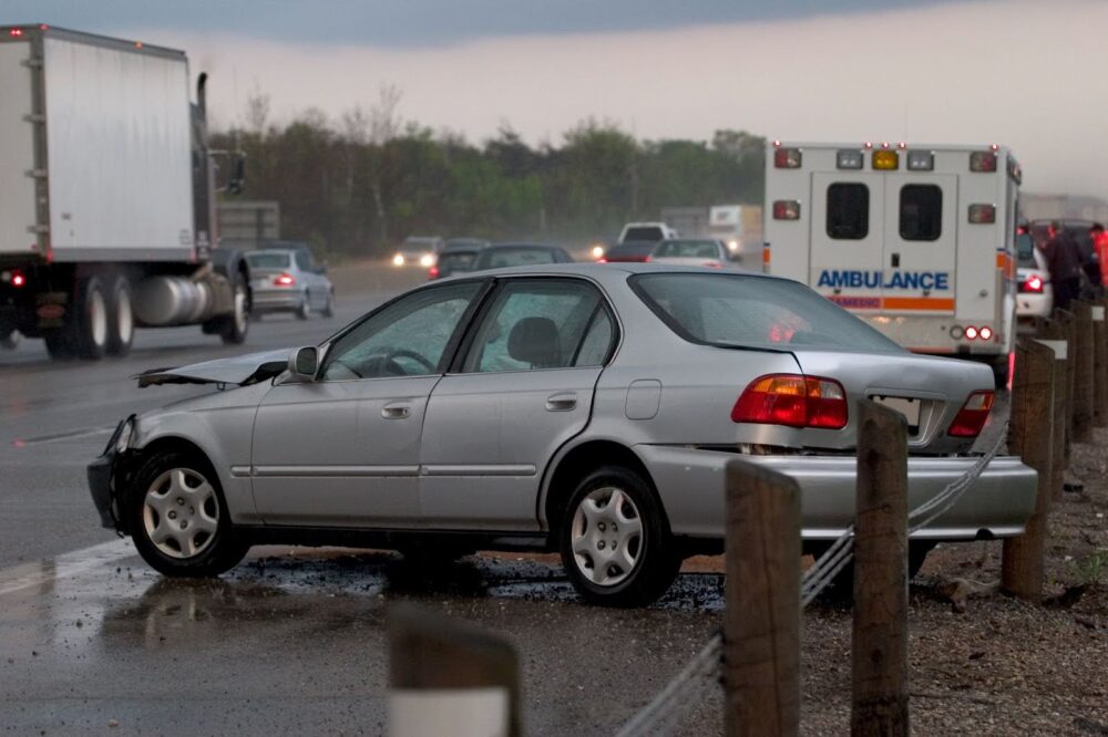 A wrecked silver car on the side of the road. The car bears visible damage to the rear bumper and engine hood. There is an ambulance and police car parked a short distance from the car.