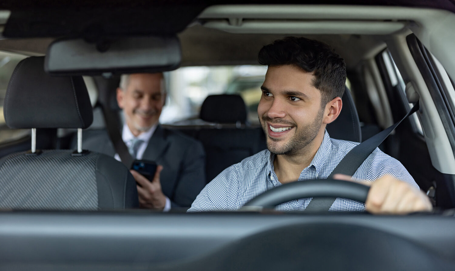 A smiling rideshare driver looks at his passenger in the rearview mirror. The passenger is smiling at his phone.