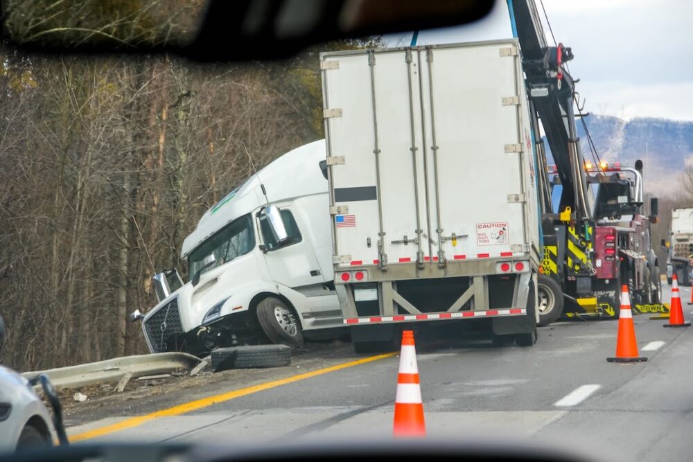 A wrecked semi-truck leaning off the side of the road. The trailer is positioned normally, but the cab is turned around to the left of the trailer at an angle. Cones are set up on the road while traffic passes by.