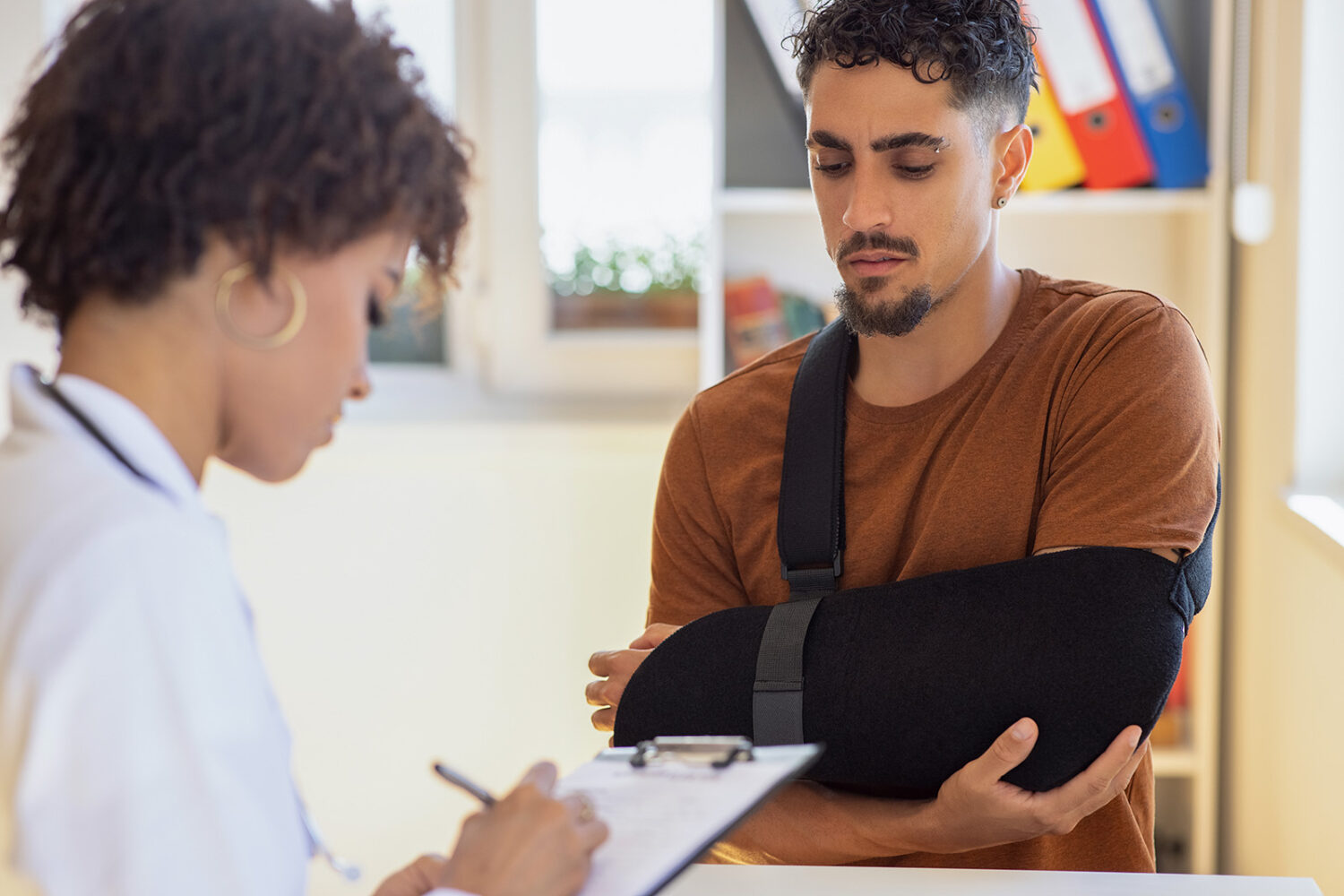 A man with his injured arm in a sling speaks with a medical provider who is taking notes on a clipboard.