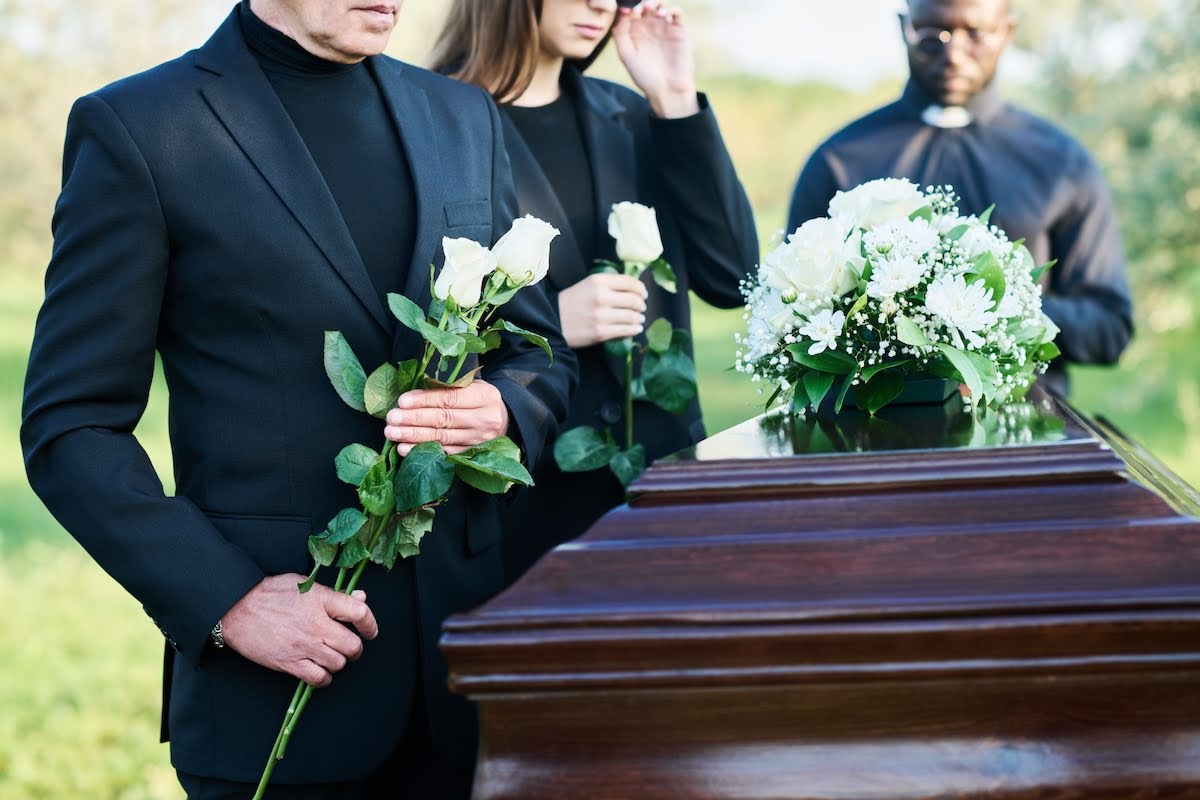 A man and woman holding white roses standing beside a dark coffin at the funeral of a loved one. A clergyman is seen behind the coffin, his head is bowed as if reading from a book.
