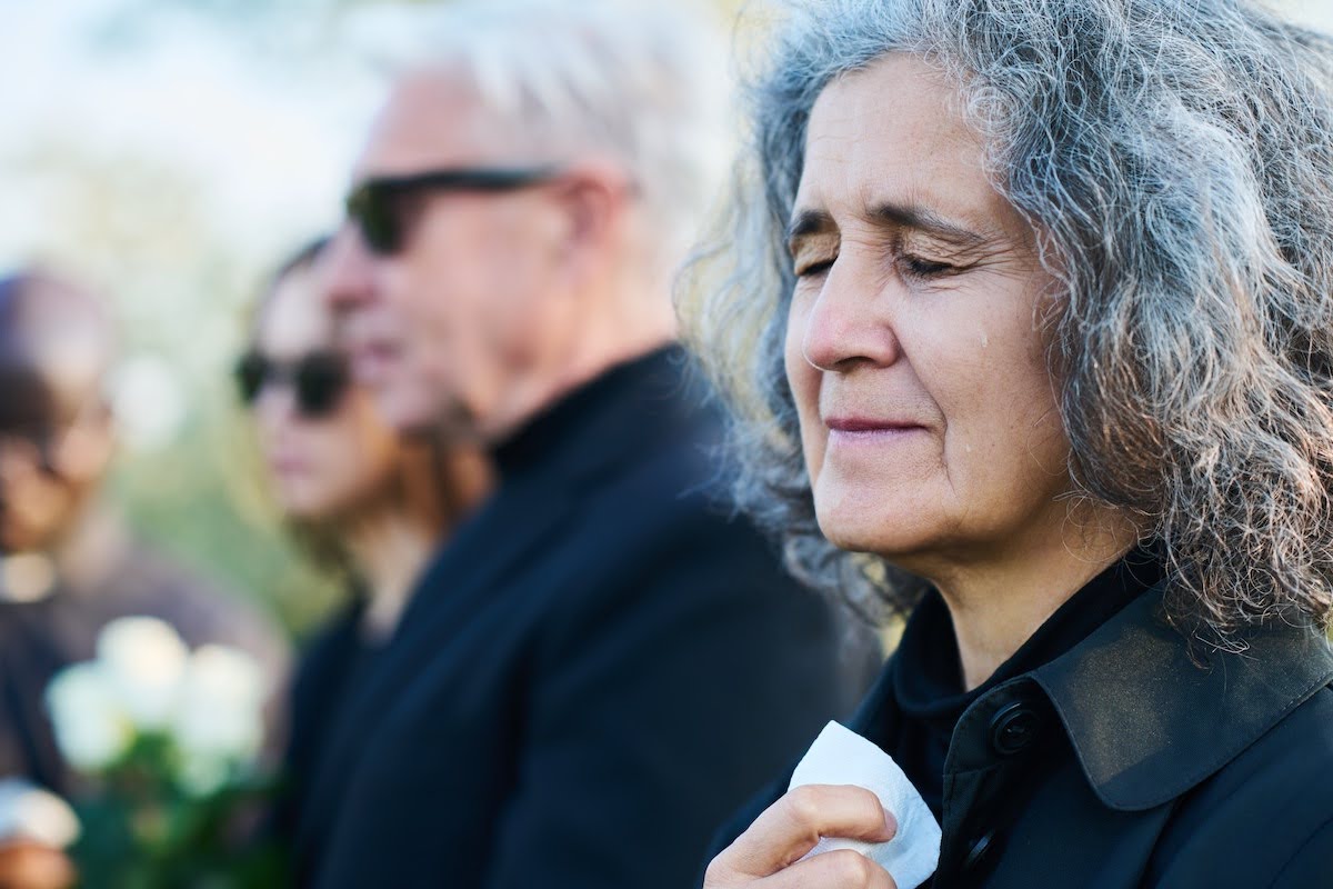 A mature woman with her eyes closed holds a handkerchief at the funeral of a loved one. Other funeral guests are seen out of focus in the background.