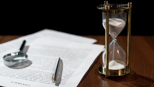 A mostly full hourglass stands on a desk next to legal documents with a magnifying glass and a pen sitting on top of the documents.