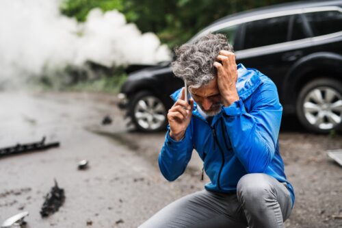 Mature man squatting in the road in front of a steaming, wrecked crossover vehicle.