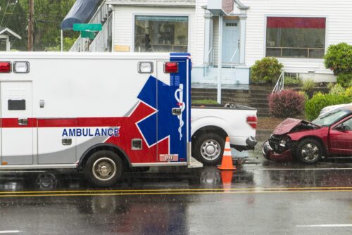 An ambulance responds to a rear-end accident on a wet roadside, where a damaged red car sits behind a white pickup truck.