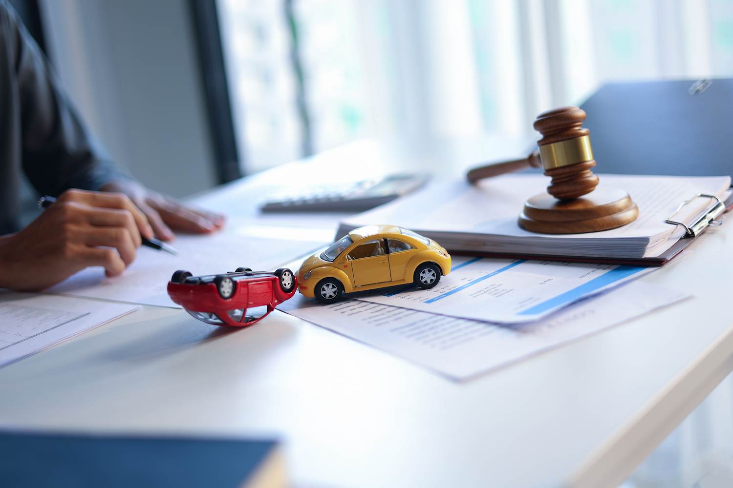 A red toy car and yellow toy car placed as if they were in an accident, sitting on a lawyer’s desk with paperwork and a gavel.