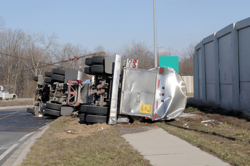 A wrecked tanker truck rolled over on its side over a sidewalk on the side of the road.