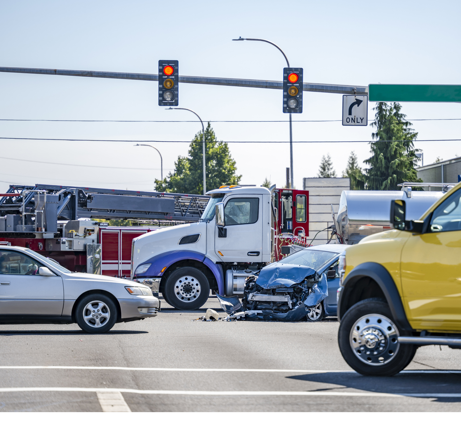 A damaged passenger car sits in an intersection after a collision with a commercial truck, surrounded by a fire truck and other vehicles.